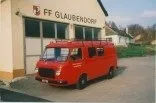 Ein roter Lieferwagen mit Gepäck auf dem Dach steht vor der FF Glaubergdorf-Feuerwehrstation. Das Gebäude hat große Fenster und ein rotes Dach.