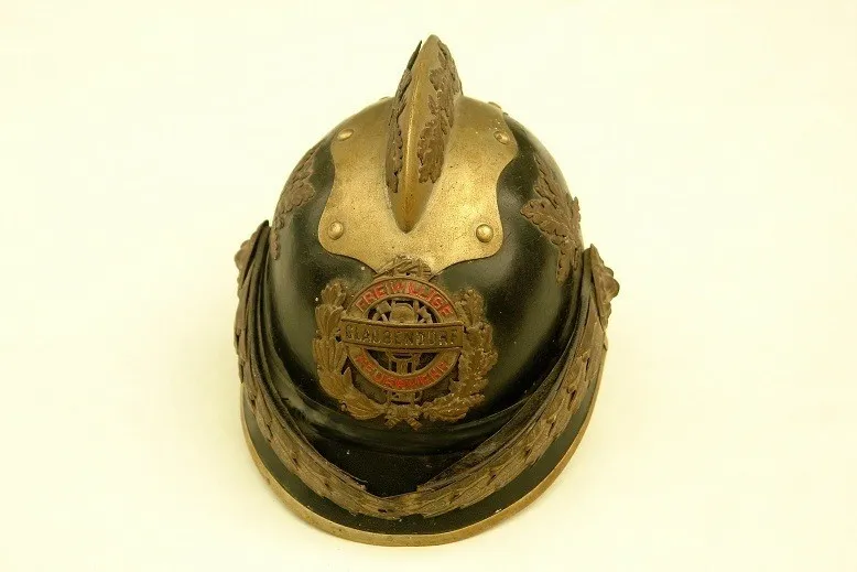 Ein altertümlicher schwarzer und goldener Helm mit verzierten Designs, der ein Messingemblem mit der Aufschrift 'BREWING' aufweist. Der Helm hat einen Metallspitz und ist auf einem weißen Hintergrund montiert.
