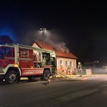 Ein Feuerwehrauto ist am Straßenrand geparkt, seine Lichter sind eingeschaltet und es spritzt Wasser auf ein brennendes Haus.