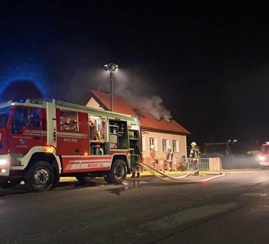 Ein Feuerwehrauto ist am Straßenrand geparkt, seine Lichter sind eingeschaltet und es spritzt Wasser auf ein brennendes Haus.