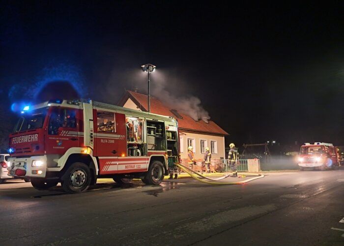 Ein Feuerwehrauto ist am Straßenrand geparkt, seine Lichter sind eingeschaltet und es spritzt Wasser auf ein brennendes Haus.