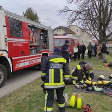 Feuerwehrleute in Uniform arbeiten auf dem Boden in der Nähe eines Feuerwehrwagens. Hinter ihnen stehen und beobachten Leute. Bäume und Gebäude sind im Hintergrund zu sehen.