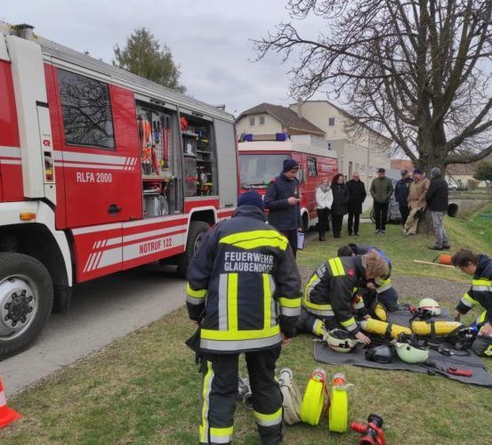 Feuerwehrleute in Uniform arbeiten auf dem Boden in der Nähe eines Feuerwehrwagens. Hinter ihnen stehen und beobachten Leute. Bäume und Gebäude sind im Hintergrund zu sehen.