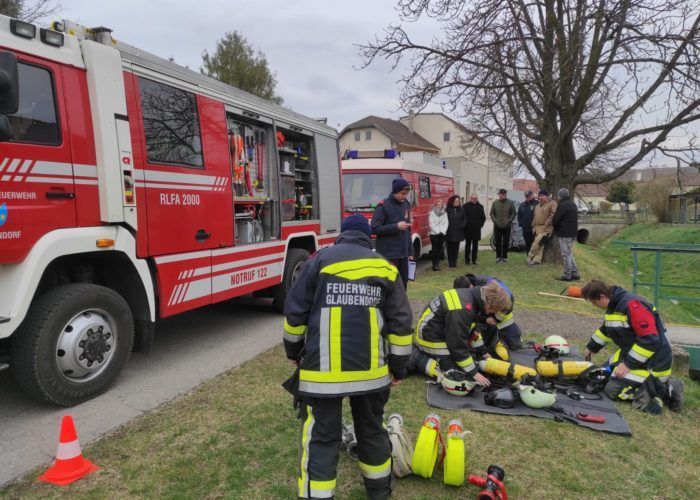 Feuerwehrleute in Uniform arbeiten auf dem Boden in der Nähe eines Feuerwehrwagens. Hinter ihnen stehen und beobachten Leute. Bäume und Gebäude sind im Hintergrund zu sehen.