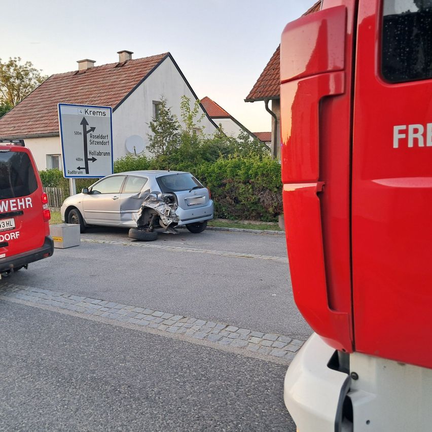 Ein silberner Wagen steht am Straßenrand mit einem platten Reifen. Dahinter weist ein Schild auf Rosendorf hin. Ein Feuerwehrwagen ist in der Nähe geparkt.