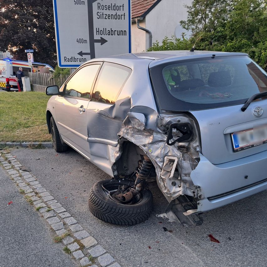 Unfallstelle mit einem stark beschädigten silbernen Auto, einem Platten und einem beschädigten Heck. Ein Schild mit Entfernungen zu Roseldorf und Hollabrunn ist sichtbar.