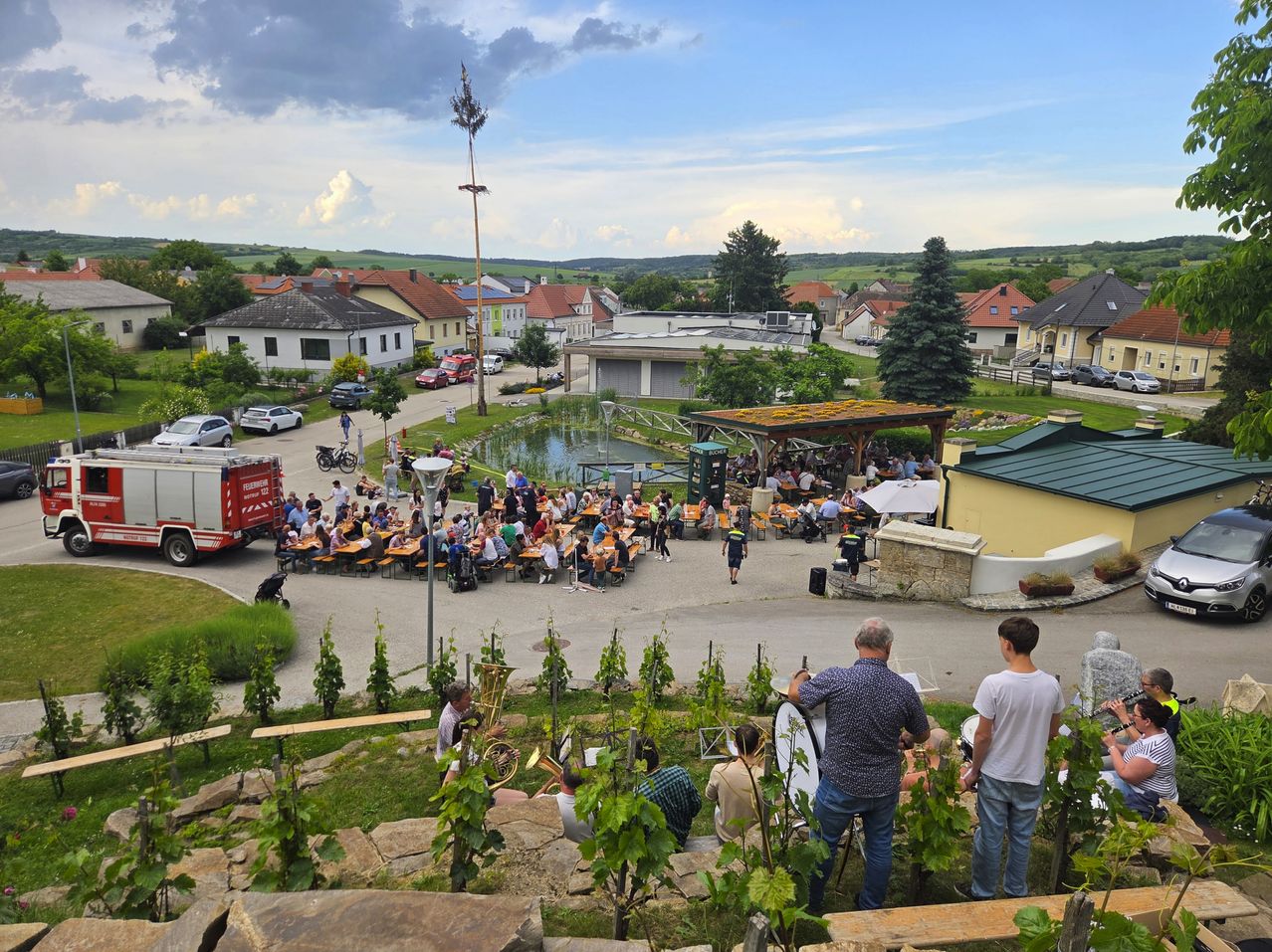 Eine Versammlung von Menschen bei einer Outdoor-Veranstaltung mit aufgestellten Tischen und Stühlen. Einige spielen Instrumente, während andere sitzen. Es gibt Häuser und einen Feuerwehrwagen in der Nähe.