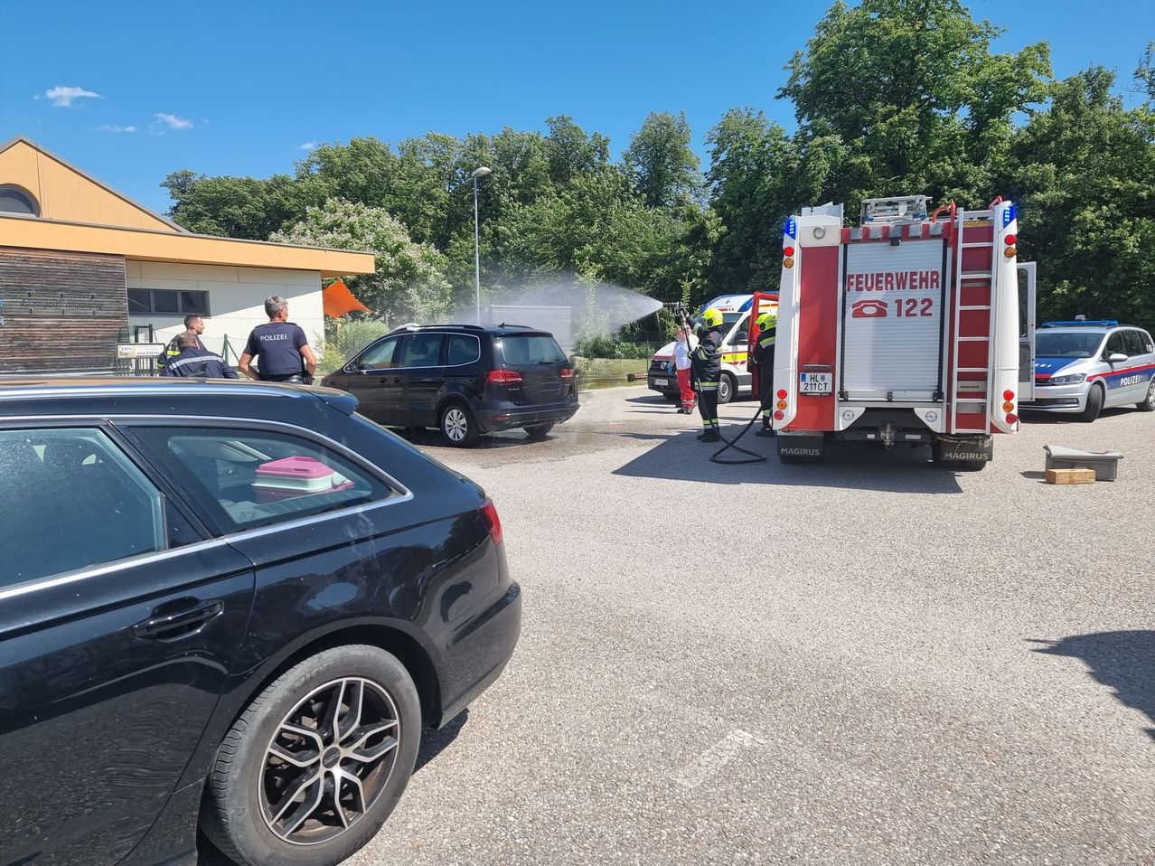 Ein Feuerwehrauto und ein Polizeiauto stehen in der Nähe eines Gebäudes. Zwei Feuerwehrleute sprühen Wasser. Im Hintergrund befinden sich Bäume und eine Straßenlaterne.