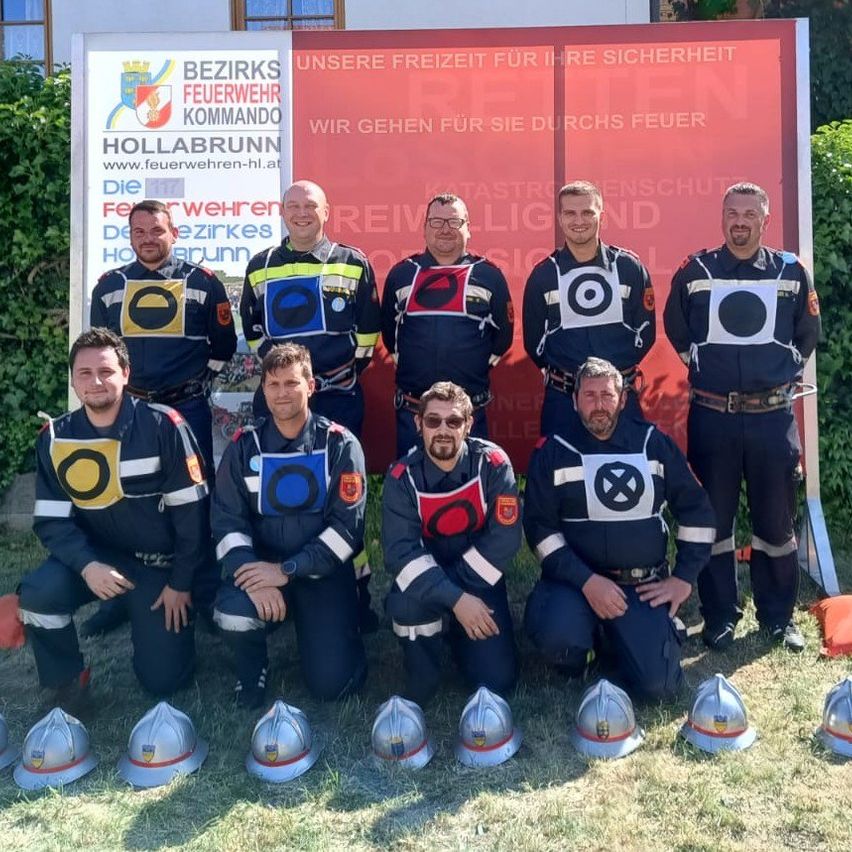 Eine Gruppe von Feuerwehrleuten in Uniform steht vor einem Schild und lächelt für ein Foto. Helme sind vor ihnen auf dem Gras platziert.