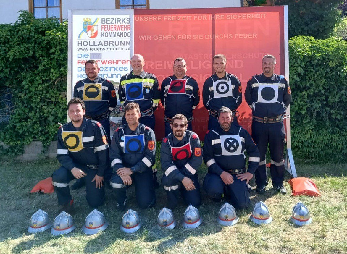 Eine Gruppe von Feuerwehrleuten in Uniform steht vor einem Schild und lächelt für ein Foto. Helme sind vor ihnen auf dem Gras platziert.