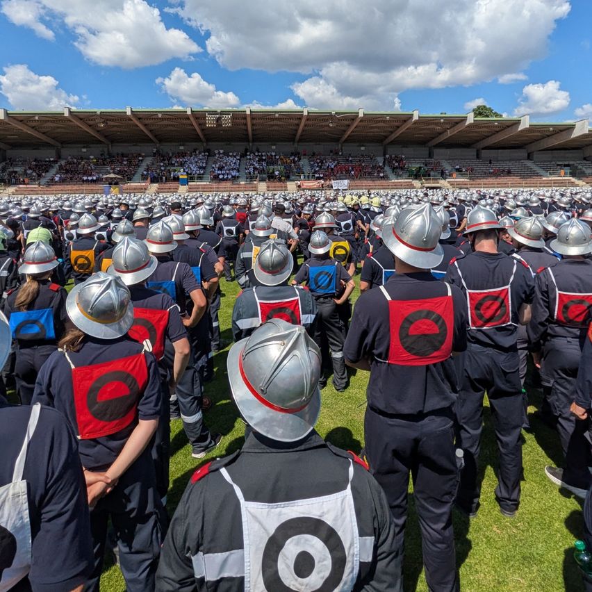 Eine große Gruppe von Personen in Sicherheitsuniformen und Helmen steht auf einem Feld vor einem Stadion unter einem bewölkten Himmel.