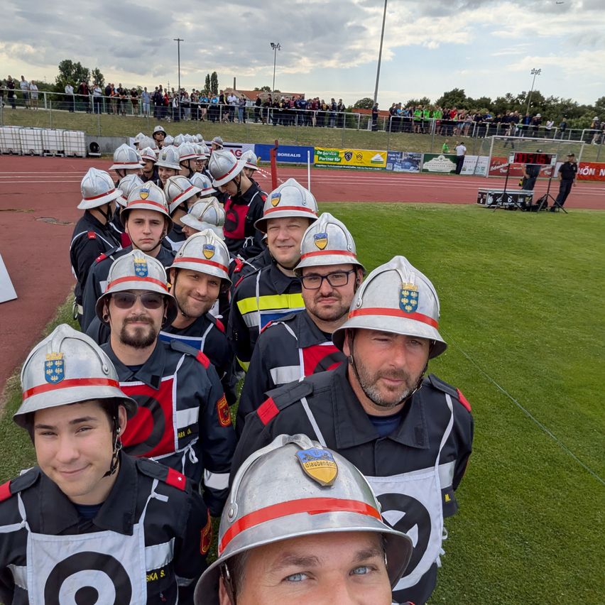 Eine Gruppe Feuerwehrleute in Uniform, einschließlich Helme, posiert für ein Foto auf einem grünen Feld mit Zuschauern im Hintergrund.