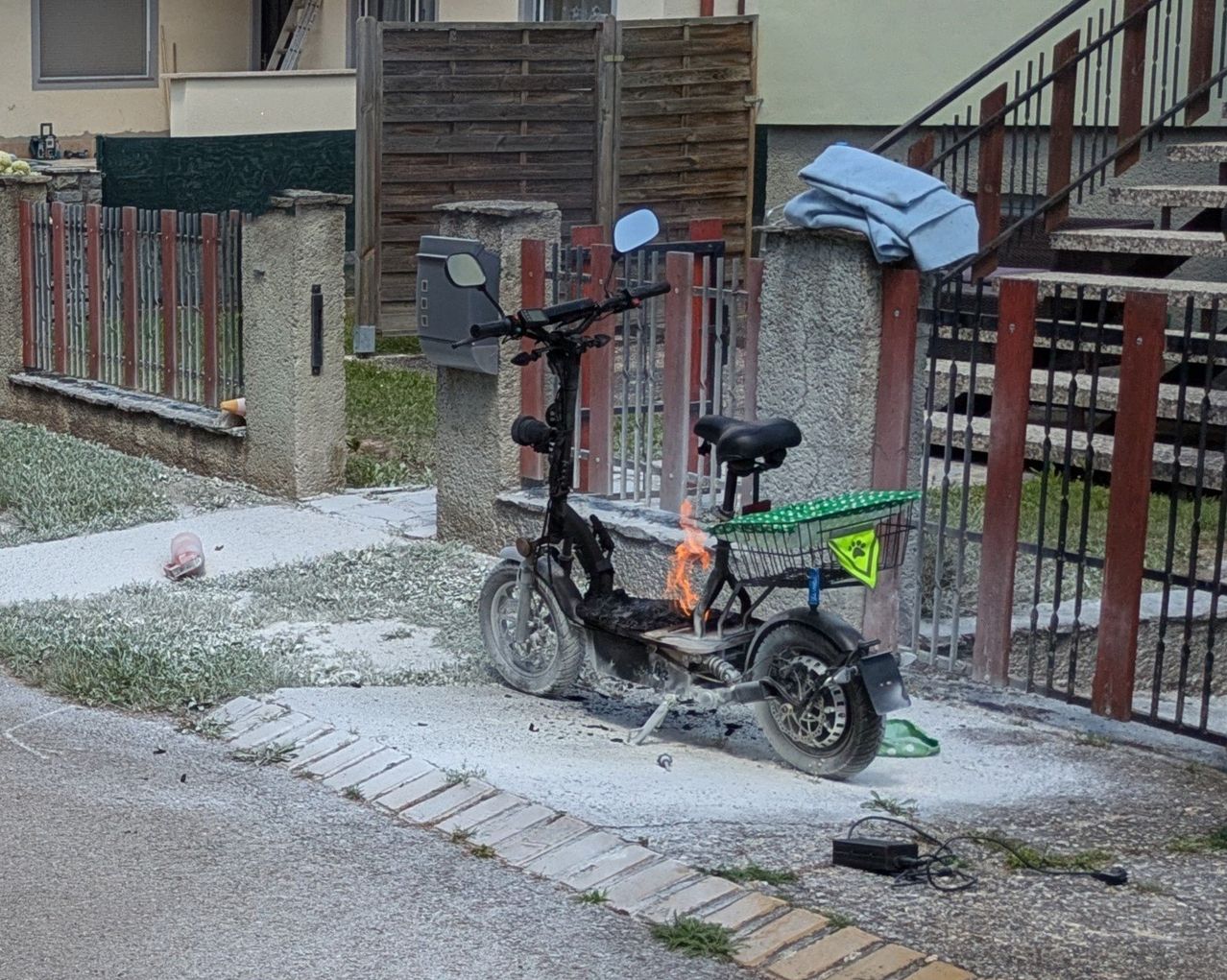 Ein ausgebrannter Elektroroller steht an einem Zaun gelehnt, mit einem Korb daran befestigt. Hinter dem Roller befindet sich ein Haus mit einer Treppe.