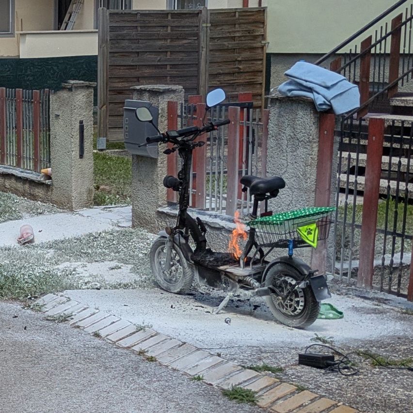 Ein ausgebrannter Elektroroller steht an einem Zaun gelehnt, mit einem Korb daran befestigt. Hinter dem Roller befindet sich ein Haus mit einer Treppe.