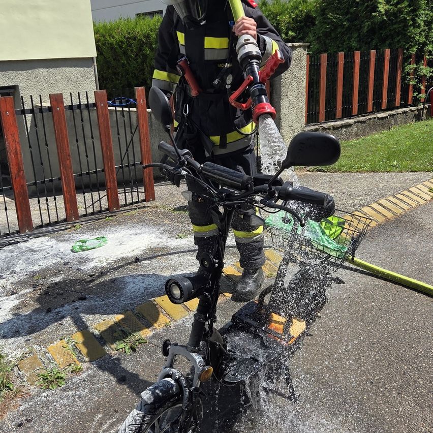 Ein Feuerwehrmann sprüht Wasser auf ein Fahrrad mit einem Schlauch und steht auf einer nassen Straße.