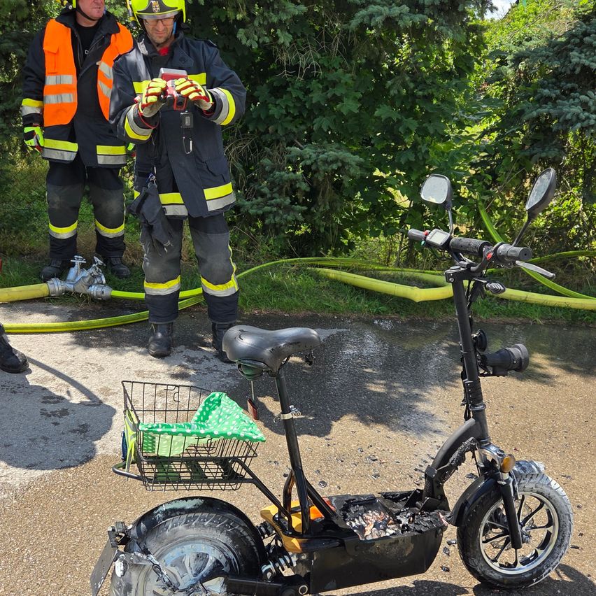 Zwei Feuerwehrleute untersuchen einen beschädigten Elektroroller mit einem Korb, einer hält eine Taschenlampe. In der Nähe liegt ein Feuerwehrschlauch auf dem Boden.