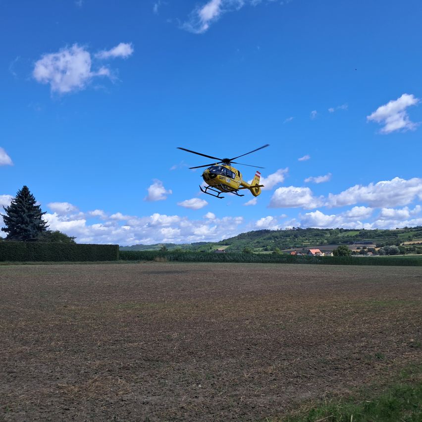 Ein gelber Hubschrauber fliegt über einem kahlen Feld bei blauem Himmel mit verstreuten Wolken. Der Boden ist braun und im Vordergrund befindet sich ein Hecken.