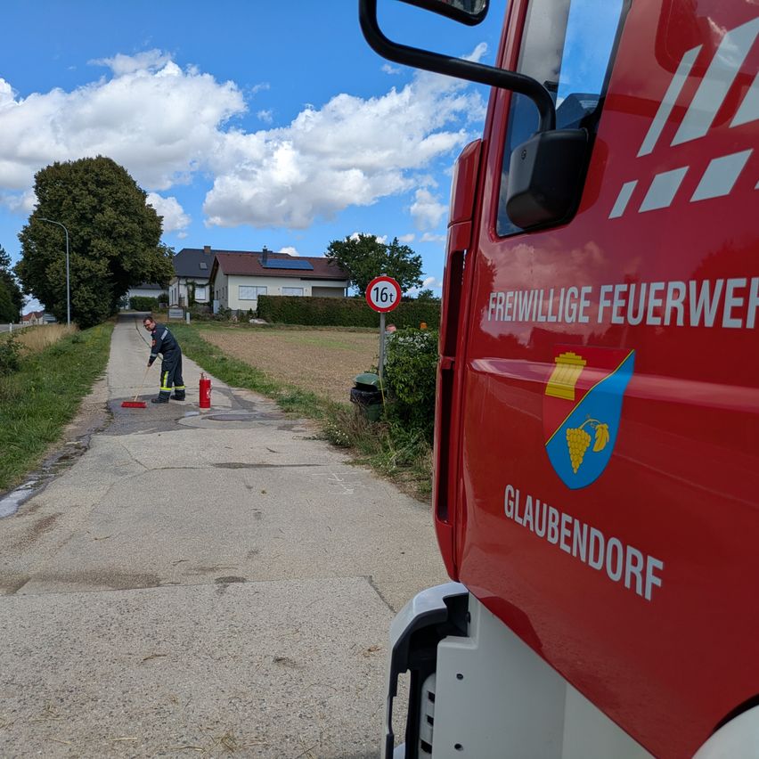 Ein freiwilliger Feuerwehrmann reinigt einen Hydranten auf einem Betonweg mit einem Haus und Solarmodulen im Hintergrund. Ein roter Feuerwehrwagen ist in der Nähe geparkt.