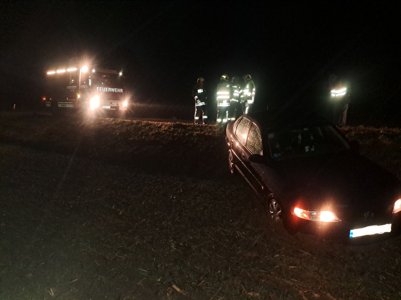 Feuerwehrleute in reflektierenden Westen stehen bei einem Unfallort in der Nacht. Ein Feuerwehrauto ist in der Nähe geparkt und beleuchtet.