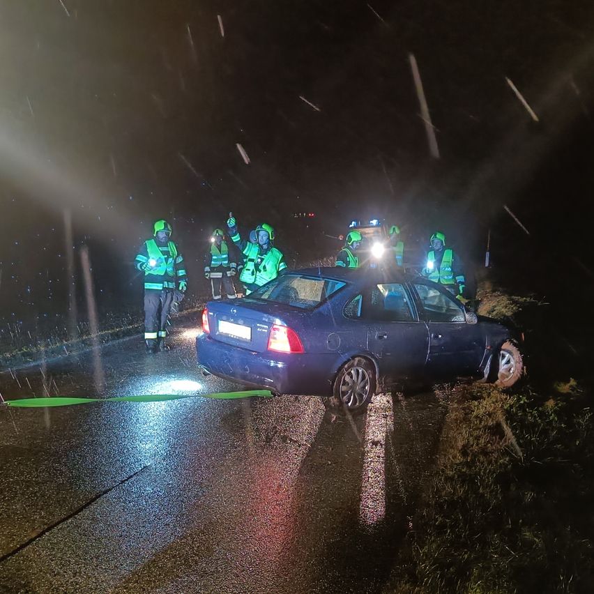 Ein blaues Auto steht mit eingeschaltetem Warnblinklicht am Straßenrand. Fünf Personen in reflektierenden Westen stehen um das Auto herum. Es ist Nacht und es regnet.