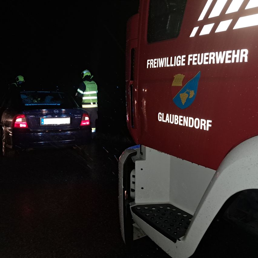 Ein Feuerwehrfahrzeug der Freiwilligen Feuerwehr Glaubendorf steht bei Nacht neben einem blauen Auto. Zwei Personen in reflektierenden Westen stehen in der Nähe.