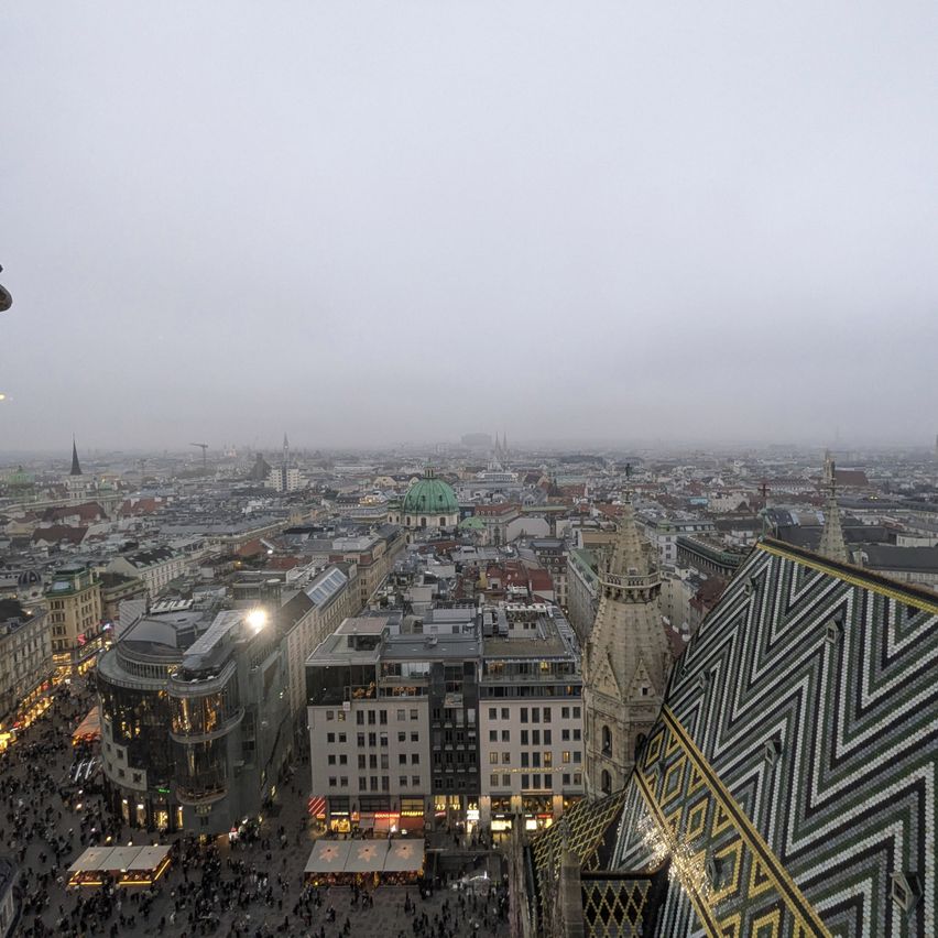 Luftaufnahme von Wien mit vielen Gebäuden, einer belebten Straße und einem bewölkten Himmel. Der berühmte Stephansdom ist sichtbar.