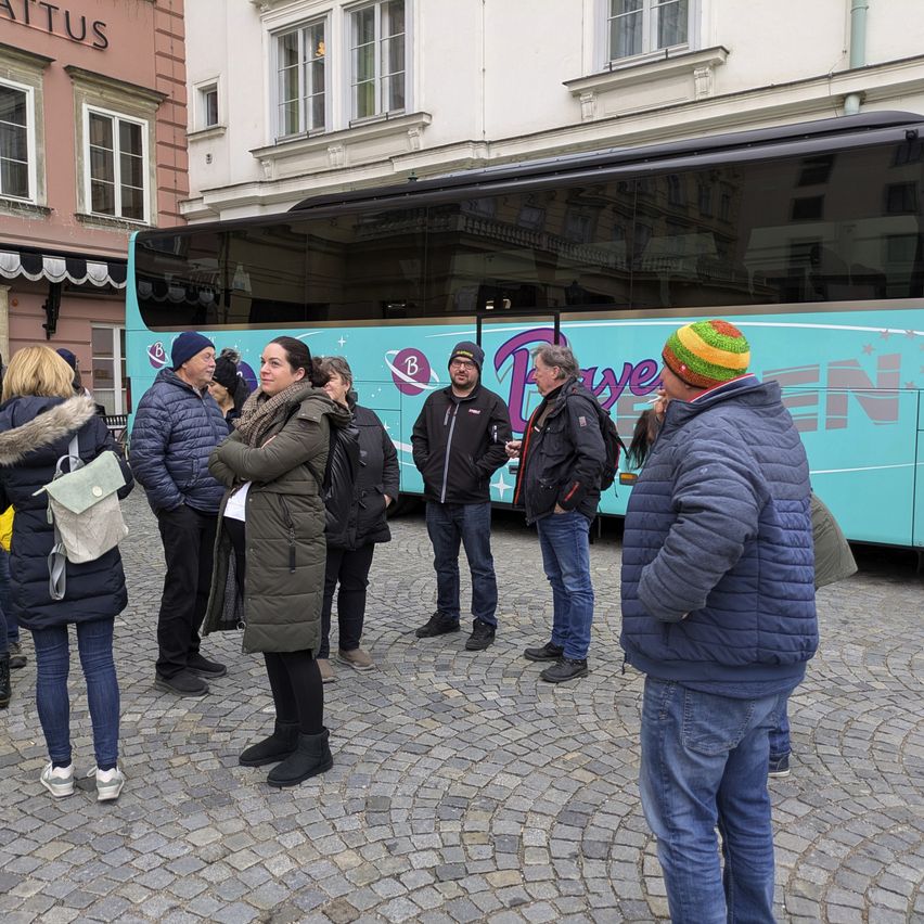 Eine Gruppe von Menschen steht vor einem Bus auf einer Kopfsteinpflasterstraße. Der Bus hat bunte Designs. Einige Leute tragen Mützen.