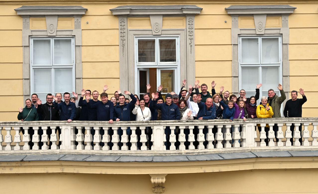 Eine Gruppe von Menschen steht auf einem Balkon vor einem Gebäude und winkt. Einige tragen Jacken und Mützen, und einige haben Handtaschen dabei. Hinter ihnen hat das Gebäude gelbe Wände, Glasfenster und dekorative Steinarbeiten.
