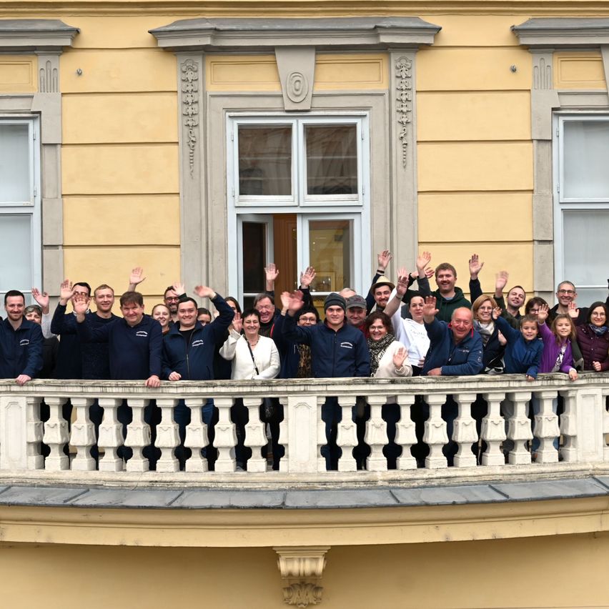Eine Gruppe von Menschen steht auf einem Balkon vor einem Gebäude und winkt. Einige tragen Jacken und Mützen, und einige haben Handtaschen dabei. Hinter ihnen hat das Gebäude gelbe Wände, Glasfenster und dekorative Steinarbeiten.