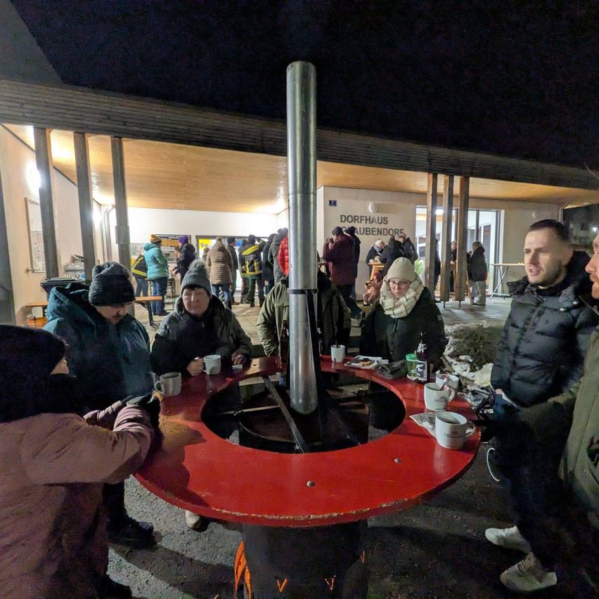 Mehrere Personen sitzen um einen runden Tisch mit Tassen und einem Schornstein in der Mitte. Es ist Nacht, und sie scheinen Getränke zu genießen. Einige Leute stehen hinter ihnen. Ein Schild sagt Dorfhaus Gaubendorf.
