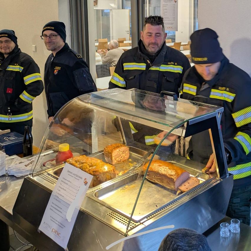Vier Feuerwehrleute stehen neben einer Vitrine mit Fleisch. Zwei von ihnen servieren das Fleisch, während die anderen beiden in der Nähe stehen.
