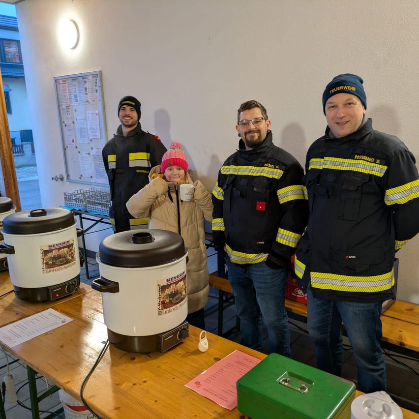 Drei Feuerwehrleute und ein lächelndes Mädchen mit einer Tasse stehen hinter einem Tisch mit zwei Reiskochern und einer grünen Box.