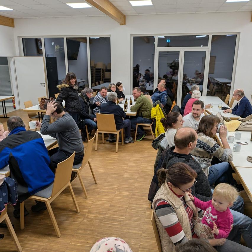 Ein Essbereich mit mehreren Erwachsenen und Kindern, die um Tische herum sitzen, einige mit Getränken und Essen. Der Raum hat große Fenster und Holzböden. Eine Frau in Schwarz steht neben einem Tisch mit einer Handtasche.