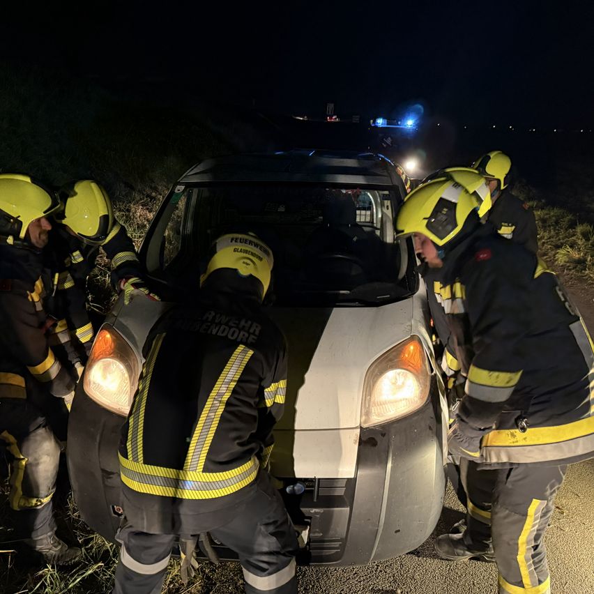 Feuerwehrleute in gelben Helmen und Schutzkleidung arbeiten an einem beschädigten Fahrzeug am Straßenrand bei Nacht.