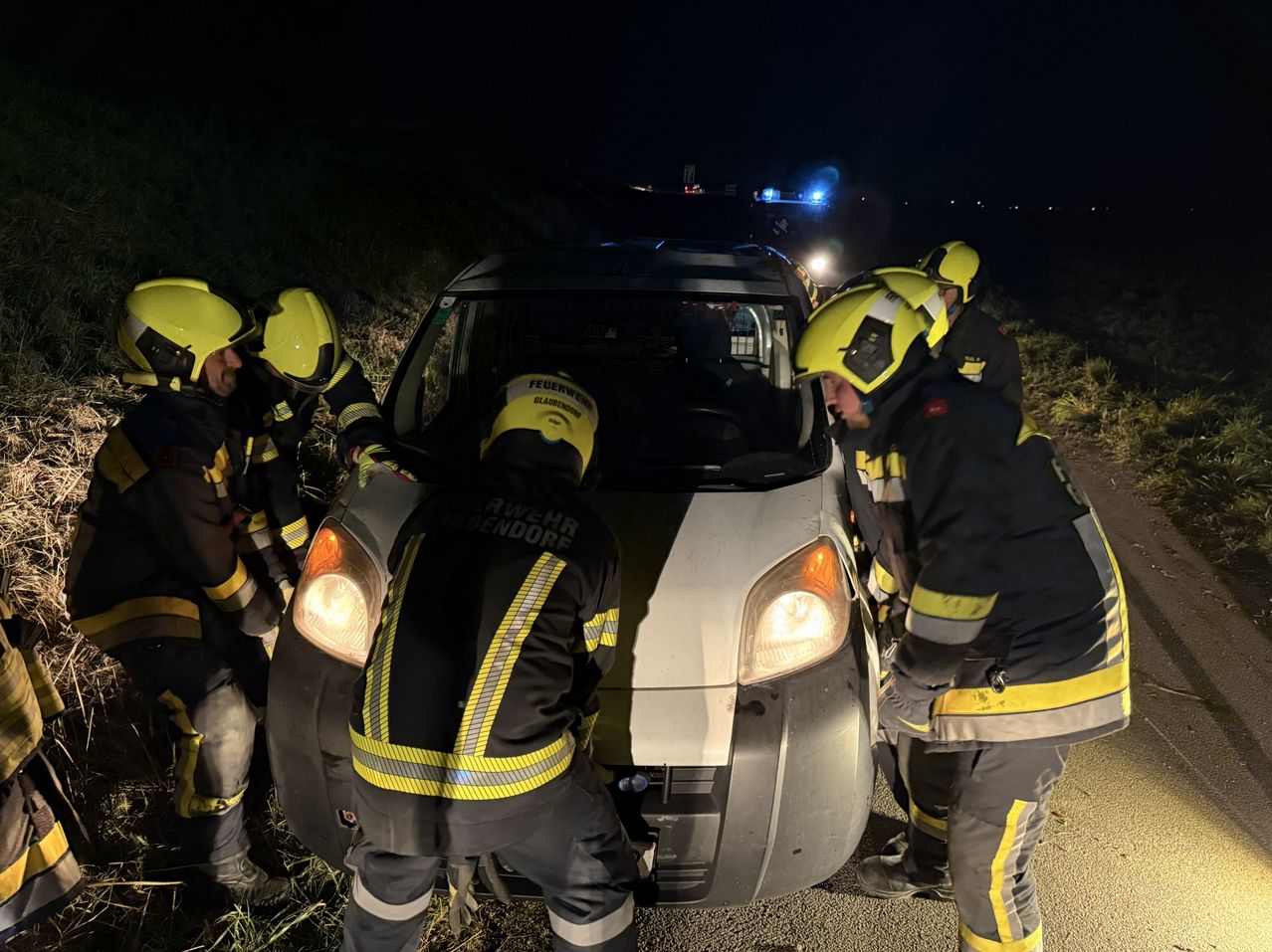Feuerwehrleute in gelben Helmen und Schutzkleidung arbeiten an einem beschädigten Fahrzeug am Straßenrand bei Nacht.