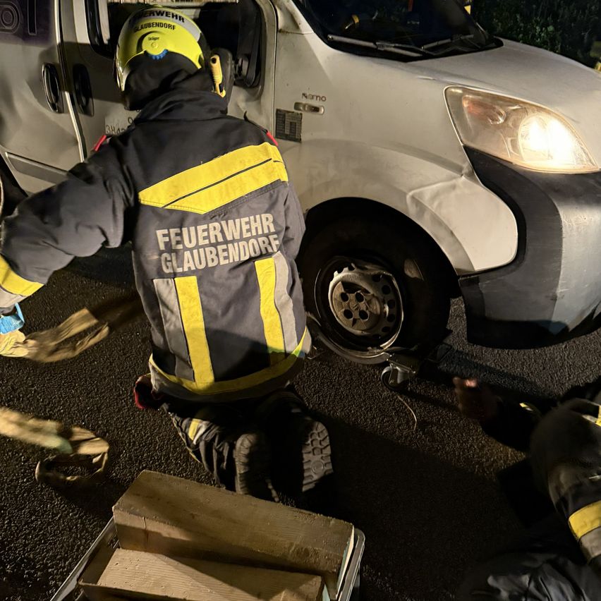 Ein Feuerwehrmann mit dem Aufkleber 'Feuerwehr' auf seiner Jacke ist zu sehen, wie er mit einer anderen Person an einem Fahrzeug arbeitet. Sie sind am Straßenrand.