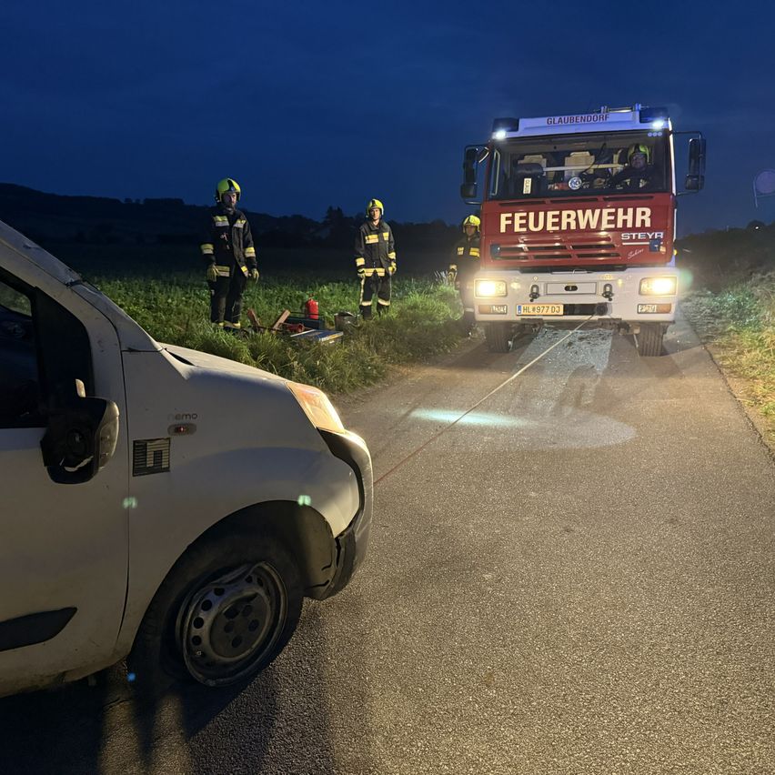 Ein weißer Van ist am Straßenrand geparkt. Ein Feuerwehrwagen mit 'FEUERWEHR' und 'STEYR' Markierungen steht in der Nähe, seine Scheinwerfer sind eingeschaltet. Feuerwehrleute sind anwesend, einer liegt am Boden, andere stehen herum.