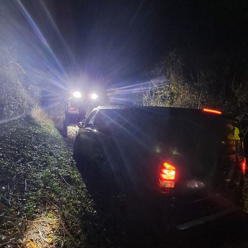 Zwei Fahrzeuge sind auf einem grasigen Hang bei Nacht geparkt. Ein Fahrzeug ist dunkel, und das andere ist ein Traktor mit eingeschalteten Scheinwerfern. Eine Person steht hinter dem Traktor.
