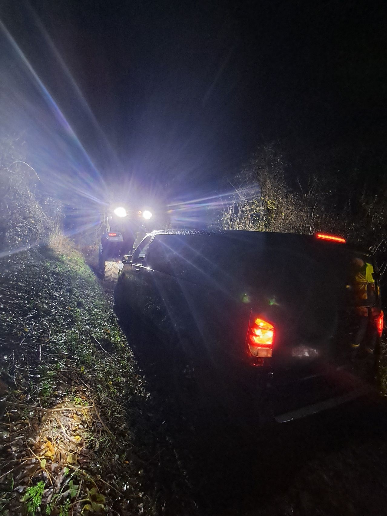 Zwei Fahrzeuge sind auf einem grasigen Hang bei Nacht geparkt. Ein Fahrzeug ist dunkel, und das andere ist ein Traktor mit eingeschalteten Scheinwerfern. Eine Person steht hinter dem Traktor.