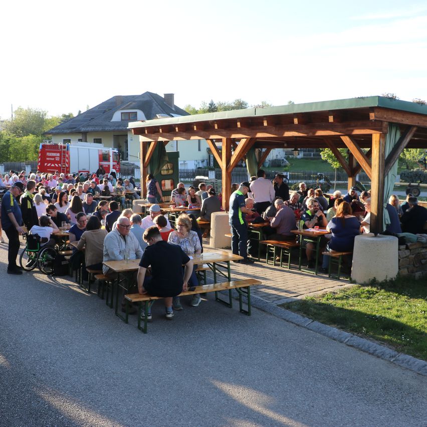 Eine Gruppe von Menschen sitzt an Picknicktischen unter einem Pavillon. Ein Feuerwehrmann steht links, und ein roter Feuerwehrwagen ist dahinter geparkt. Ein Rollstuhl ist ebenfalls vorhanden.