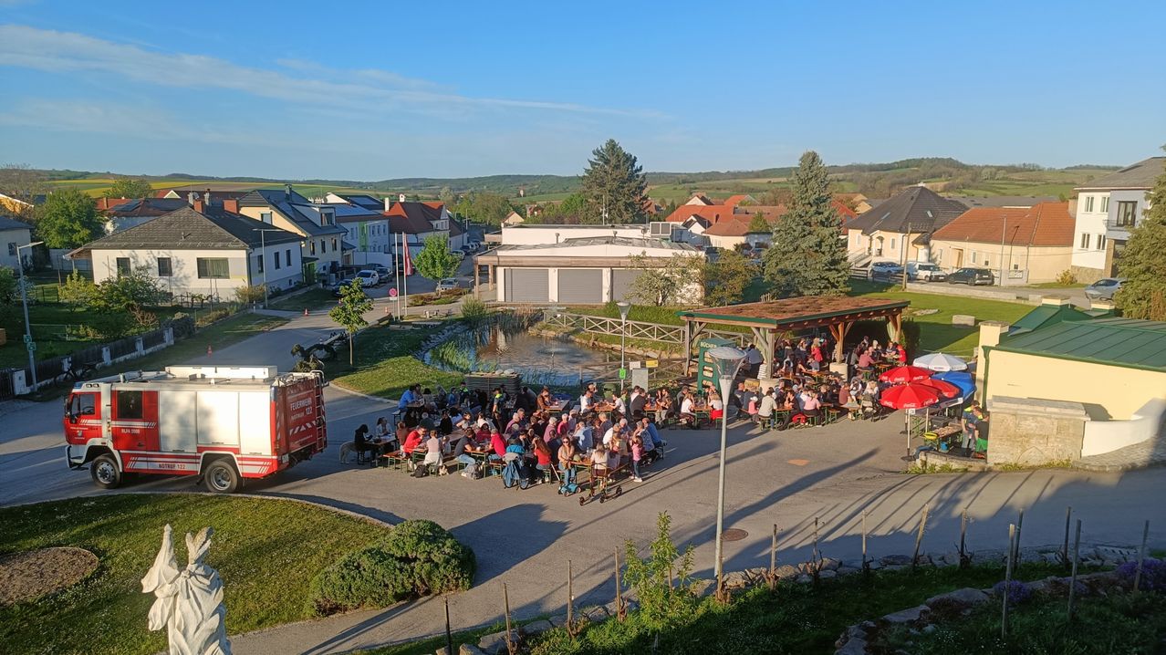 Ein Luftbild eines Dorfes zeigt Menschen, die im Freien unter Sonnenschirmen essen. Ein Feuerwehrauto ist in der Nähe geparkt.