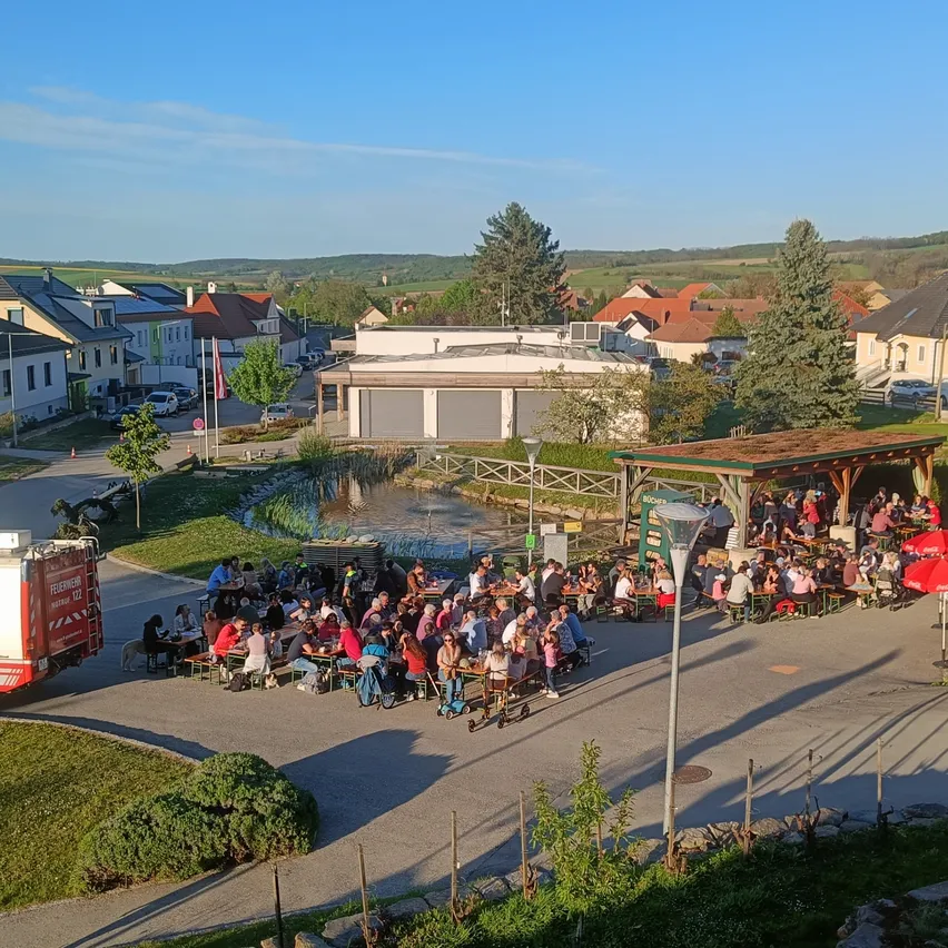 Ein Luftbild eines Dorfes zeigt Menschen, die im Freien unter Sonnenschirmen essen. Ein Feuerwehrauto ist in der Nähe geparkt.