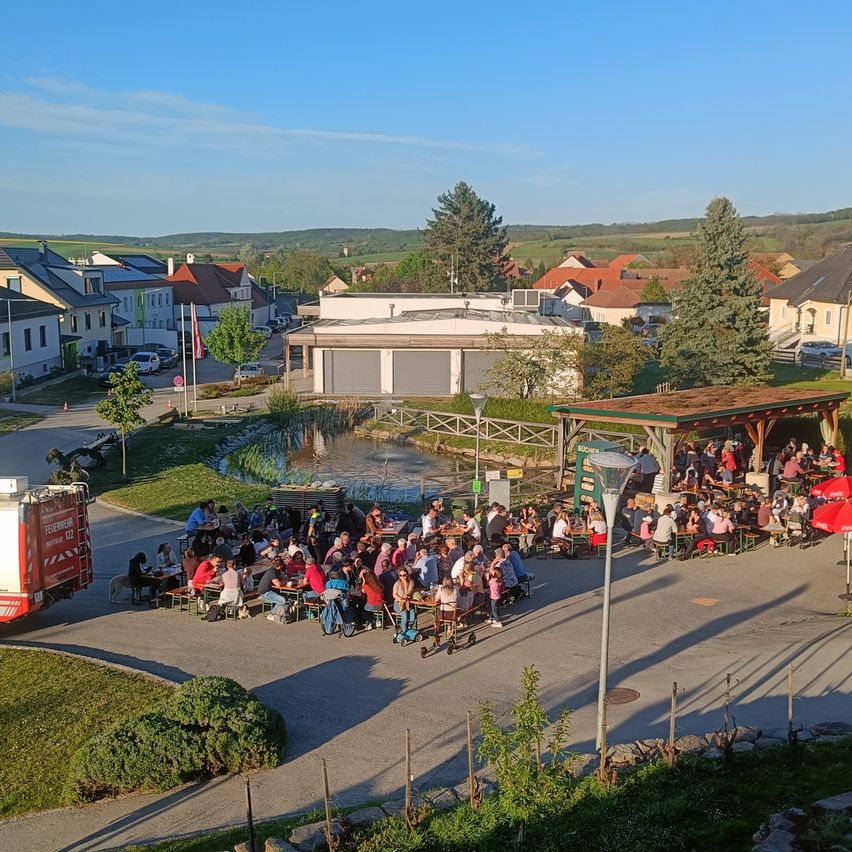 Ein Luftbild eines Dorfes zeigt Menschen, die im Freien unter Sonnenschirmen essen. Ein Feuerwehrauto ist in der Nähe geparkt.