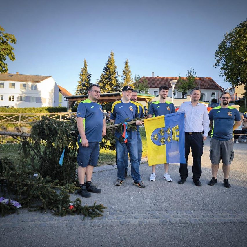 Fünf Männer in blauen Trikots halten eine Flagge und Äste. Sie stehen an einem sonnigen Tag draußen, mit Häusern und Bäumen im Hintergrund.