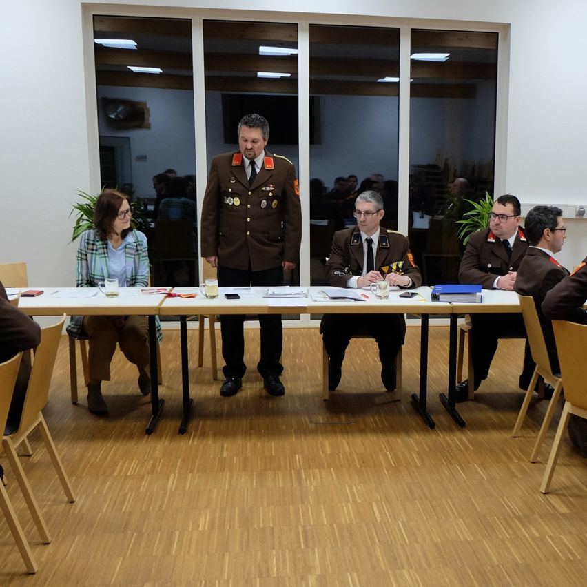 Mehrere Personen in Militäruniformen sitzen um einen Tisch in einem Besprechungsraum herum. Ein Mann steht an der Kopfseite des Tisches und spricht die Gruppe an.