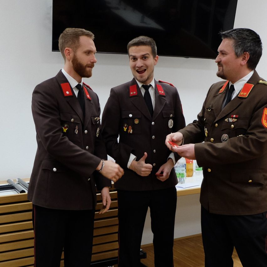 Drei Männer in Militäruniformen stehen lächelnd in einem Raum. Der Mann auf der rechten Seite hält einen kleinen Gegenstand. Hinter ihnen steht ein Schreibtisch mit Flaschen und Papieren. Ein Fernseher ist an der Wand montiert.