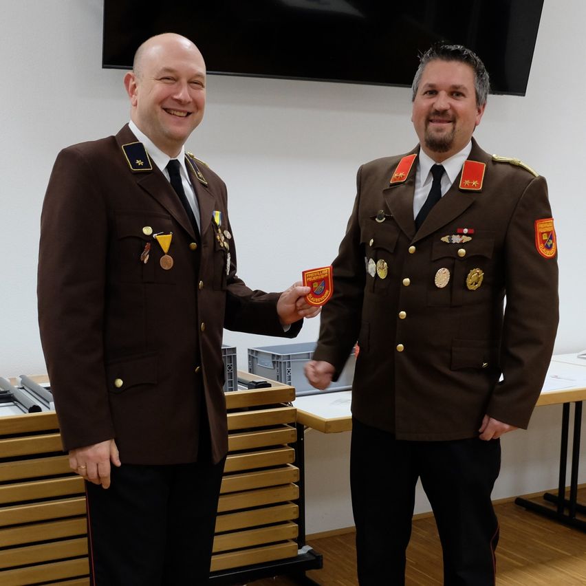 Zwei Männer in Militäruniformen stehen lächelnd da, einer überreicht dem anderen eine Medaille. Beide tragen Medaillen und Abzeichen auf ihren Uniformen. Hinter ihnen steht ein Schreibtisch mit einer Aufbewahrungsbox und ein großer Bildschirm an der Wand.
