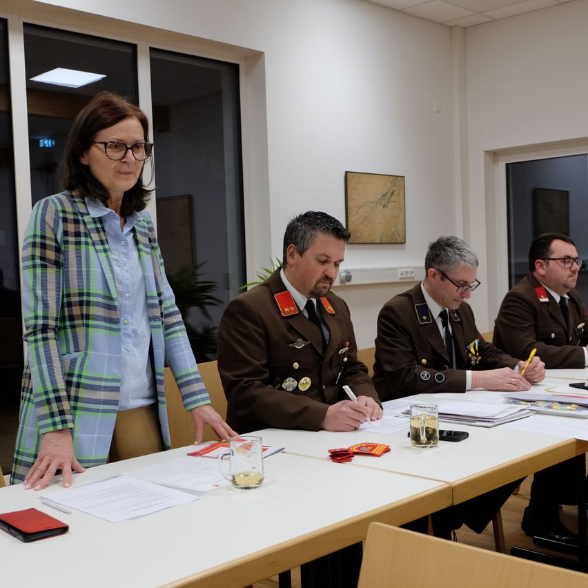 Eine Frau steht an einem Tisch bei einem Treffen mit drei Männern in Uniform, die jeweils auf Papieren schreiben. Der Tisch hat ein Glas, ein Buch und eine rote Mappe. Dahinter hält eine Wand ein gerahmtes Bild.