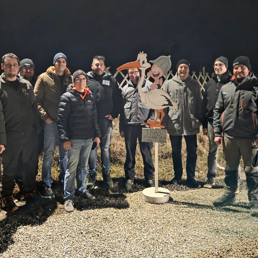 Eine Gruppe von Menschen steht lächelnd zusammen, mit einer Storchstatue in der Mitte. Sie tragen Winterkleidung und scheinen eine nächtliche Unternehmung zu unternehmen.