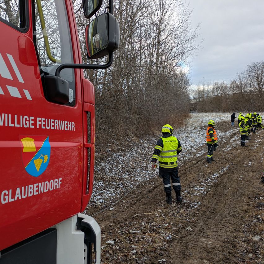 Feuerwehrleute in gelben Uniformen gehen auf einem schlammigen Pfad, mit einem roten Feuerwehrwagen im Hintergrund.